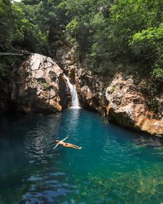 a person swimming in the water near some rocks and a waterfall with a man diving into it