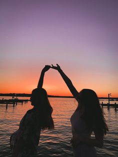 two women standing next to each other in front of a body of water at sunset