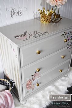 a white dresser with flowers painted on the drawers and gold handles, next to a pink flower