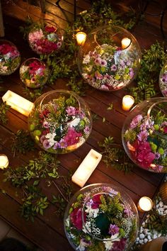 several clear bowls filled with flowers and greenery on a wooden table surrounded by candles