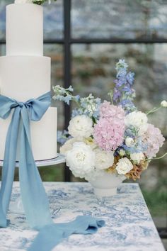 a white cake with blue ribbon and flowers in vases on a table next to a window