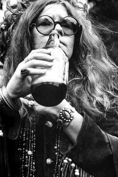 a woman with glasses drinking from a glass in front of her face and wearing beads around her neck