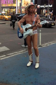 a shirtless man in shorts and cowboy boots plays guitar on the street while people walk by