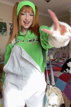 a girl with red hair wearing overalls and a green hoodie making the peace sign