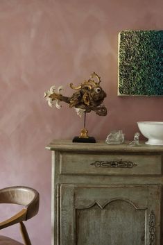 a vase with flowers on top of a wooden cabinet next to a bowl and chair