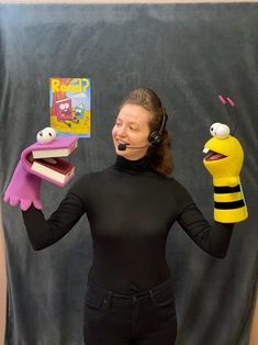 a woman wearing headphones and holding two puppets in front of her face while standing next to a black backdrop