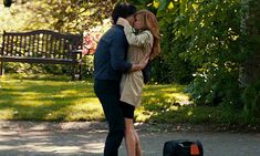 a man and woman kissing in front of a park bench with suitcases on the ground