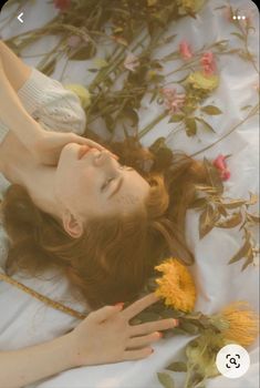 a woman laying on top of a white sheet next to yellow and pink flowers with her hands in the air