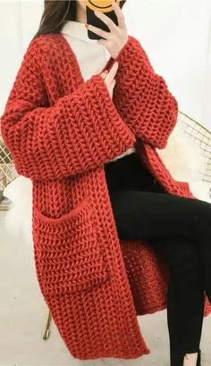 a woman sitting on a chair while holding a cell phone to her face and wearing a red knitted cardigan