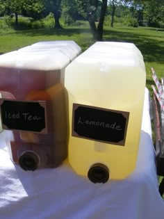 two jugs of lemonade sitting on top of a white cloth covered table with trees in the background