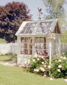 a small white garden shed sitting on top of a lush green field in front of a white picket fence