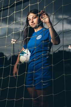 a woman holding a soccer ball in front of a goal net with dark clouds behind her