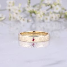 a gold ring with a red diamond in the center and flowers behind it on a white surface