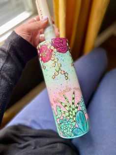 a person holding a pink and blue water bottle with flowers painted on the side, in front of a window