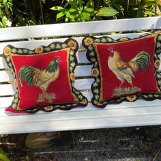 two red pillows with roosters on them sitting on a white bench in front of some bushes