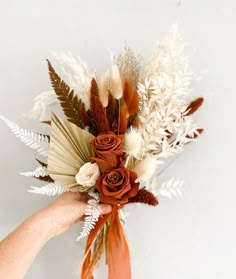 a hand holding a bouquet of flowers on top of a white wall with feathers and foliage