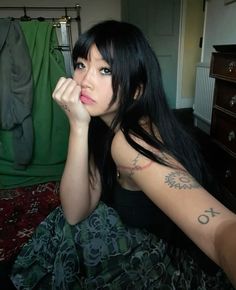 a woman with long black hair and tattoos on her arm sitting on a bed looking at the camera