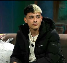 a young man sitting on top of a couch in front of a white pillow and wearing a black jacket