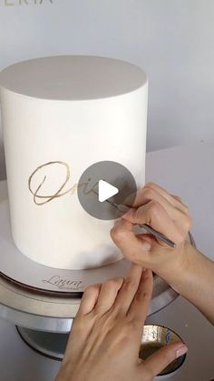 two hands are decorating a white cake with gold lettering on it and the word d is written in cursive writing