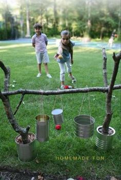 two children playing in the yard with tin cans hanging from a twig frame and some trees