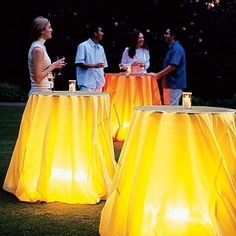 several people standing around yellow tablecloths in the grass with lights on them at night