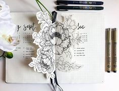 an open notebook with flowers on it next to some pens and marker markers, along with two white orchids