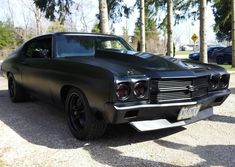 a black muscle car parked in a parking lot next to some trees and cars on the road