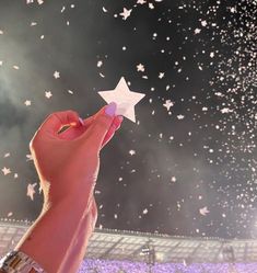 a hand holding a white paper star in front of a stadium filled with purple flowers