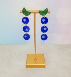 a pair of blue glass earrings with green leaves on them sitting on a gold stand