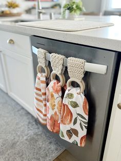 three kitchen towels hanging from hooks on a dishwasher in a kitchen with white cabinets and gray countertops