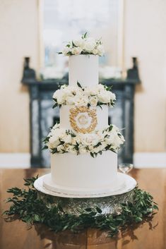 a three tiered white wedding cake with flowers on top is sitting on a table in front of a fireplace