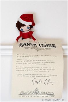 a santa claus doll is holding up a letter to santa claus on the wall behind it