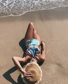 a woman laying on top of a sandy beach next to the ocean wearing a hat