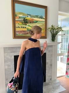 a woman in a blue dress is standing near a fireplace and holding two purses