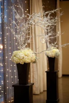 two black vases with white flowers and branches in front of a curtained window