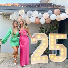 two beautiful women standing next to each other in front of balloons and the number twenty five