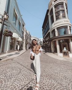 a woman standing in the middle of an empty street with buildings on both sides and people walking around