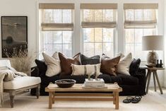 a living room filled with lots of furniture and pillows on top of a table in front of two windows