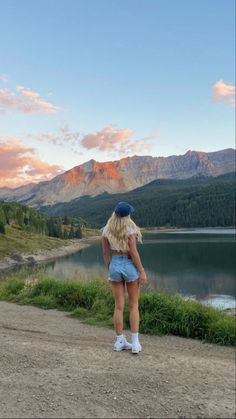 a woman standing on top of a dirt road next to a lake with mountains in the background