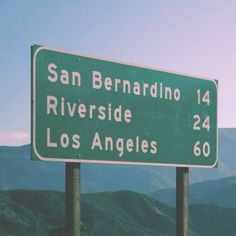 a green and white street sign sitting on the side of a road in front of mountains