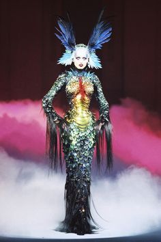 a woman in a costume with feathers on her head and long black hair, standing against a pink background