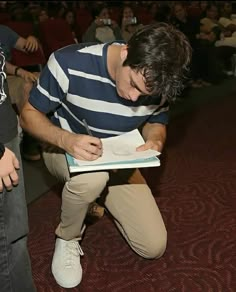 a man sitting on the floor writing in a book while people watch from behind him