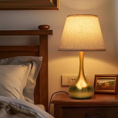 a lamp on a night stand next to a bed with a framed picture in the corner