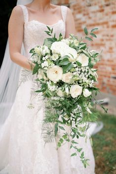 a bride holding a bouquet of white flowers and greenery on her wedding day in front of a brick wall