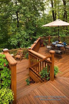 a wooden deck with chairs and an umbrella in the middle surrounded by greenery on both sides