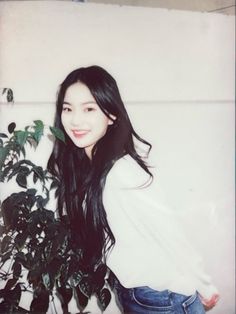 a woman standing next to a potted plant with long black hair and white shirt