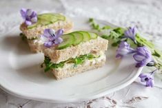 two sandwiches with cucumbers and herbs on a white plate next to purple flowers