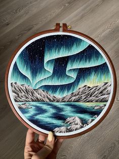 someone is holding up an embroidery project with the aurora lights in the sky above them