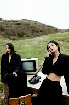 two women sitting at a desk in front of an old computer monitor and telephone, one talking on the phone