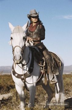 a woman riding on the back of a white horse in an open field with mountains in the background